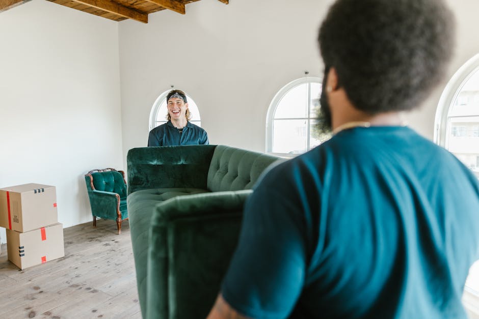 A scene inside a residential property showing two movers during a home relocation, with one mover in the foreground wearing a blue shirt and dark curly hair, holding a large green upholstered sofa. Th