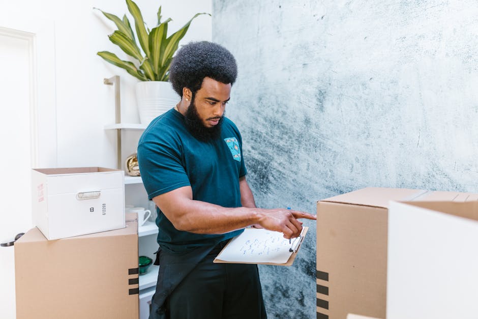 A woman sits on the floor inside a room, surrounded by large cardboard boxes used for house removals and furniture transport. She is smiling and tossing packing foam peanuts into the air, with some in